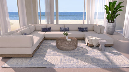 Blue 4' x 6' rug in a coastal living room, with a beige sectional and navy pillows, round wicker coffee table, sea view through glass walls, and a potted plant.