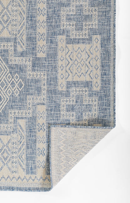 Blue and ivory geometric patchwork rug, woven fringe edges, boho kilim style, with folded corner revealing light underside.