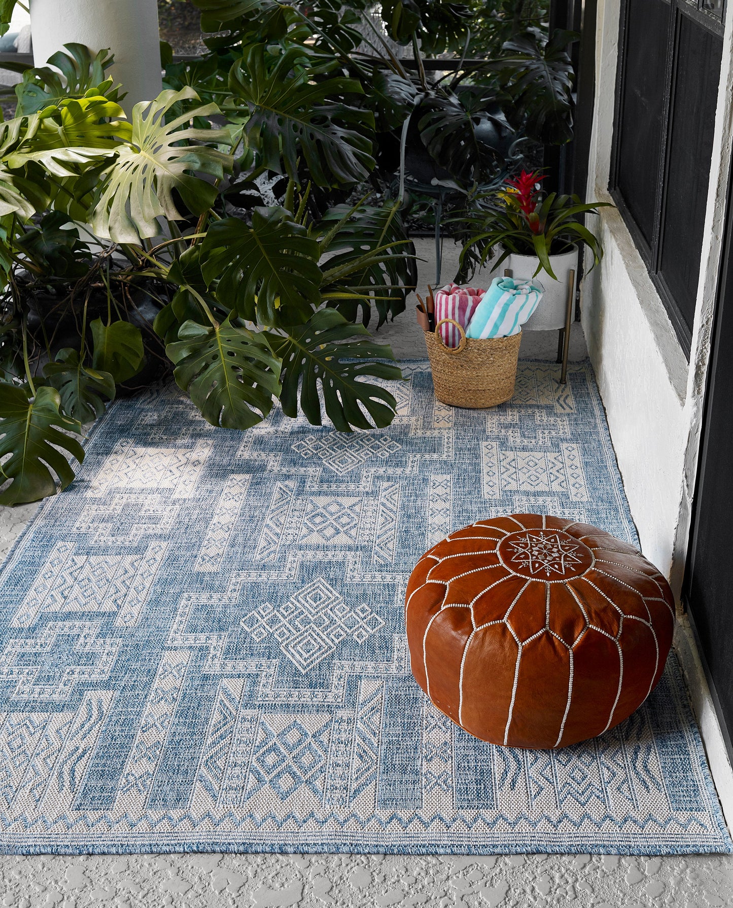 Blue geometric patterned rug on a patio with a round brown leather pouf and lush monstera plants.