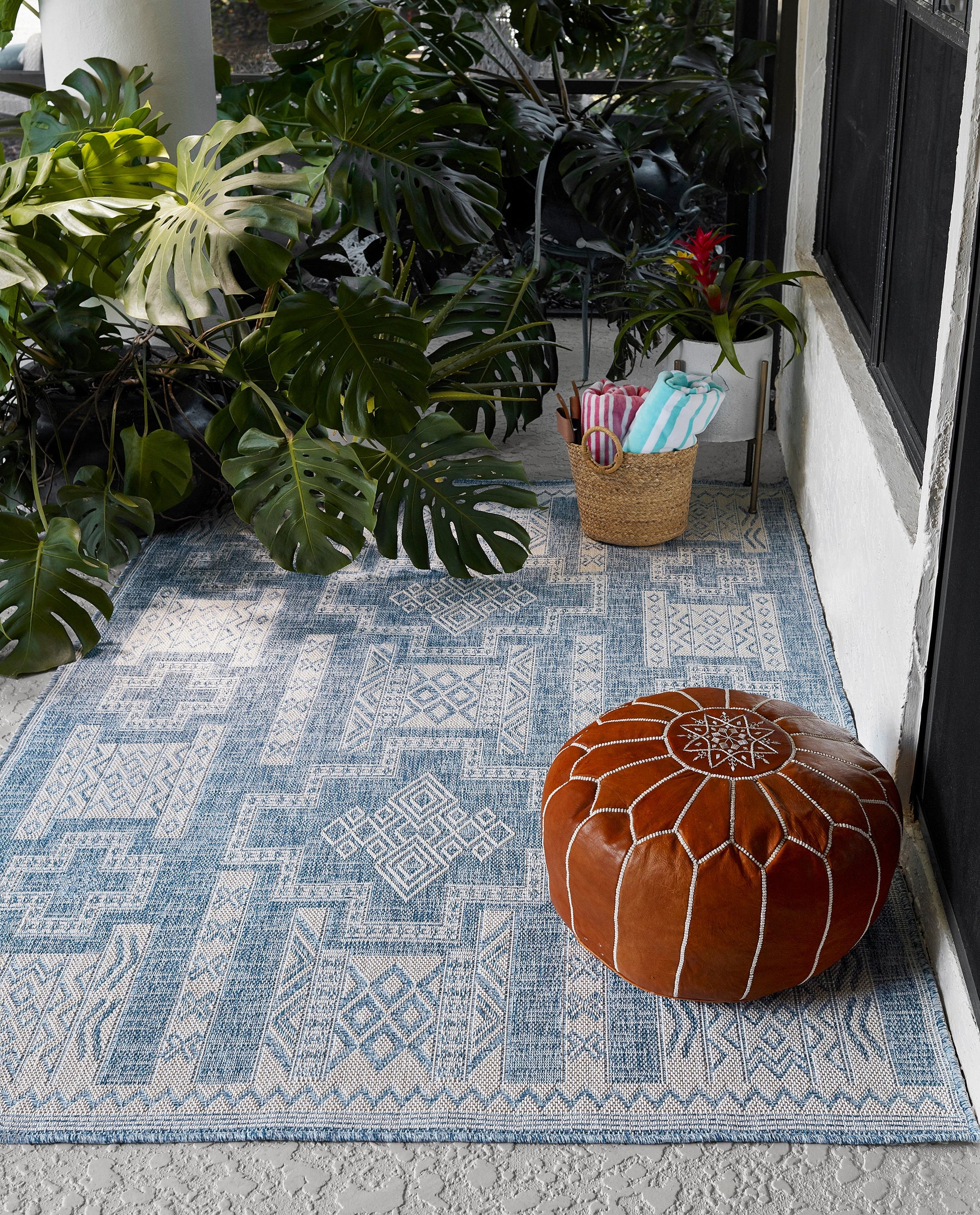 Blue patterned outdoor rug on concrete with a brown leather pouf and towel basket beside tropical plants.