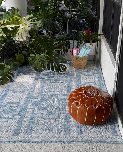 Blue patterned outdoor rug on concrete with a brown leather pouf and towel basket beside tropical plants.