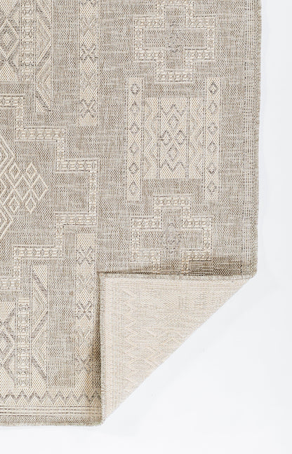 Beige-gray geometric patchwork area rug with woven texture and bordered edges; folded corner reveals lighter underside.