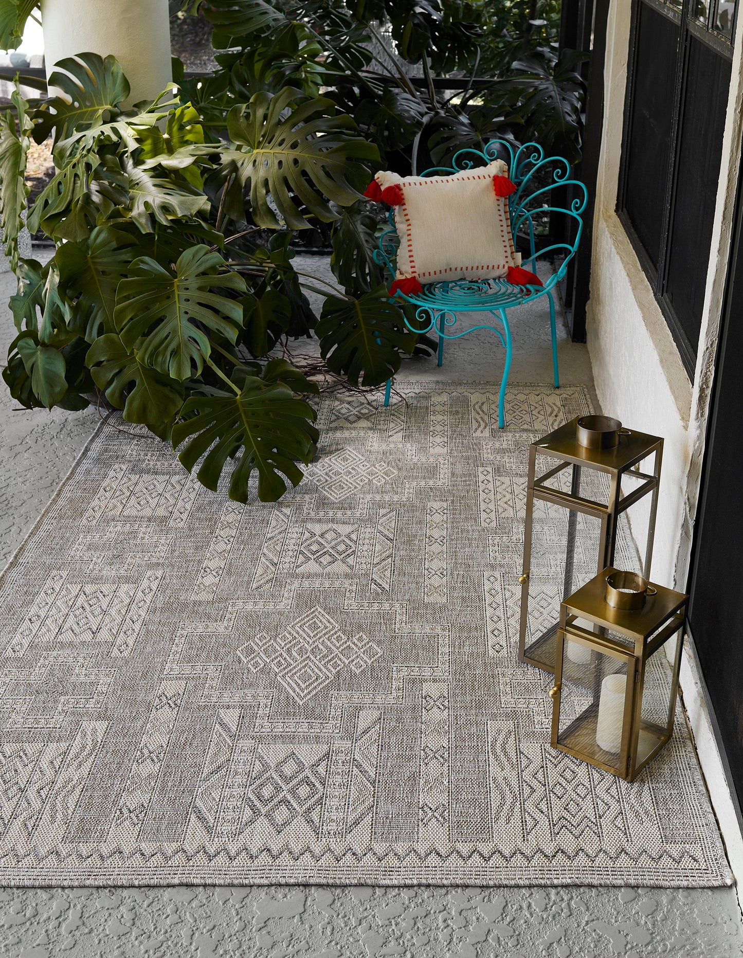 Gray geometric outdoor rug on concrete porch with turquoise metal chair, beige pillow, monstera plants, and brass lanterns.