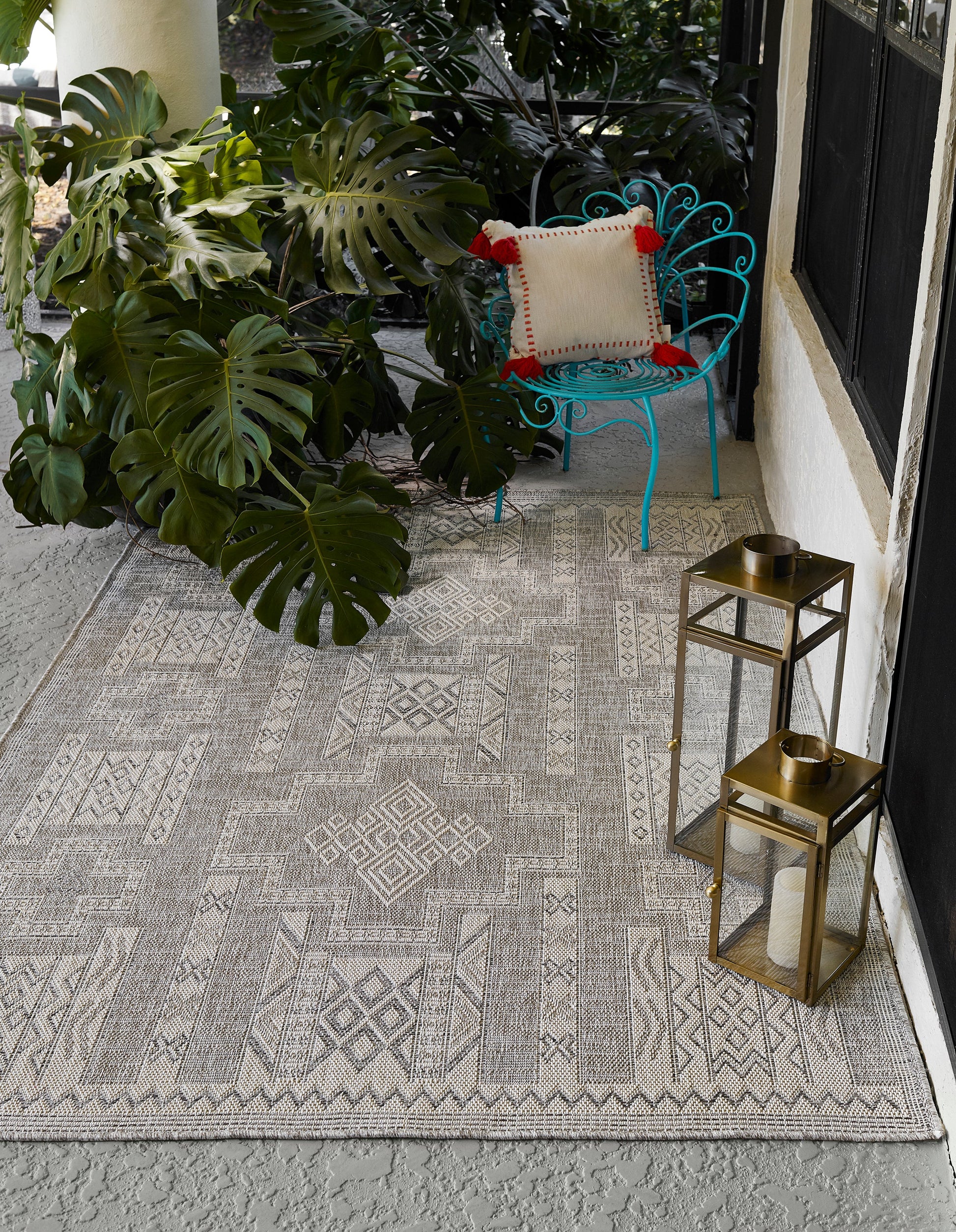 Gray geometric outdoor rug on concrete porch with turquoise metal chair, beige pillow, monstera plants, and brass lanterns.