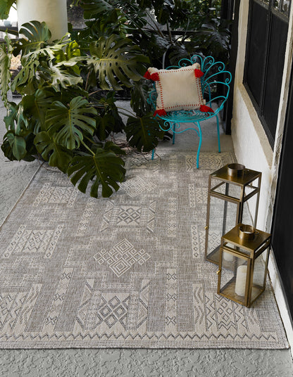 Gray geometric outdoor rug on concrete porch with turquoise metal chair, beige pillow, monstera plants, and brass lanterns.