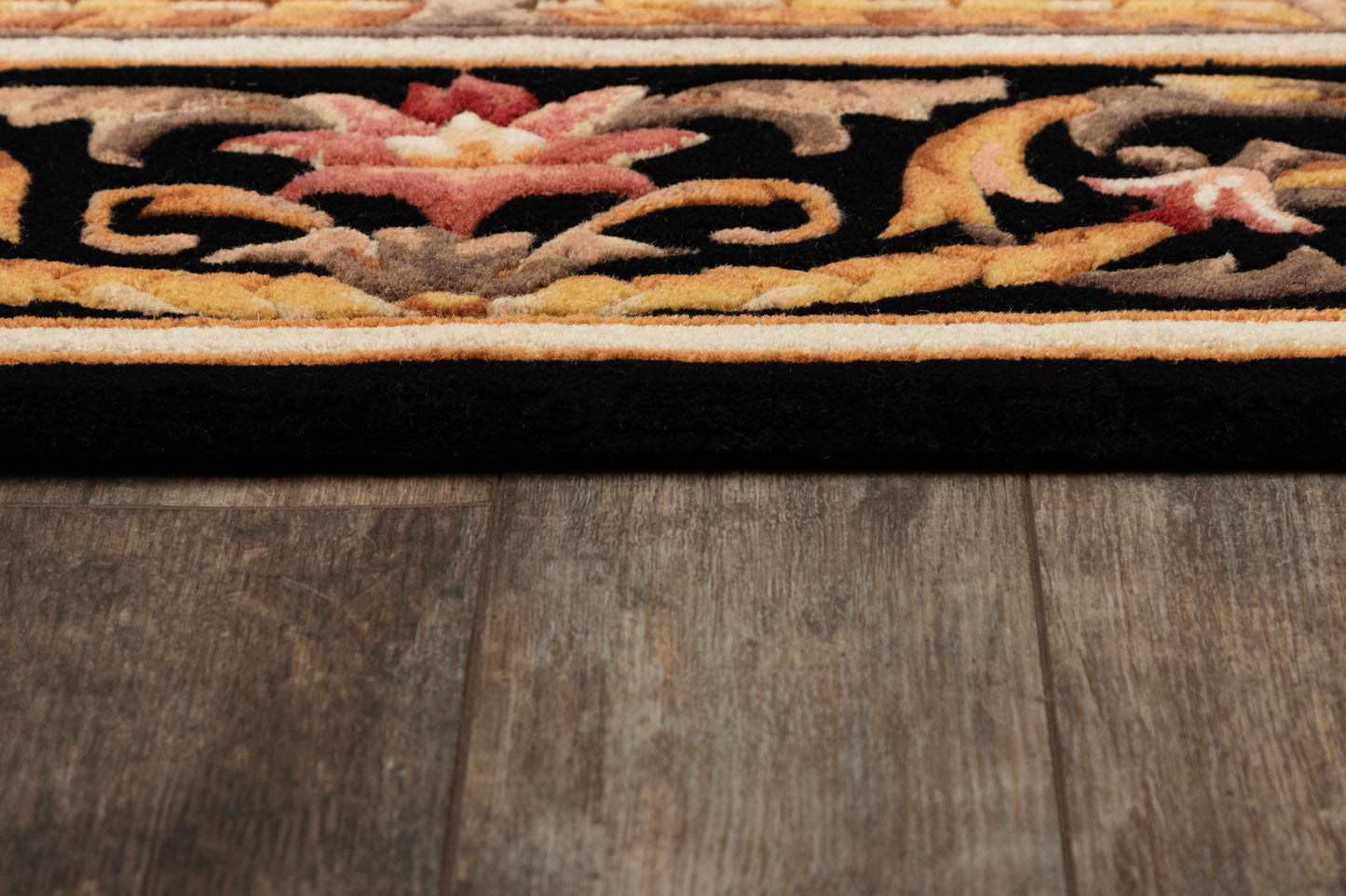 Ornate area rug edge with black field, warm orange/gold border and pink floral motif on wood floor.