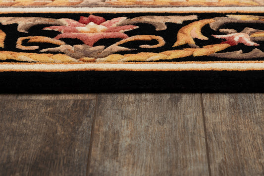 Close-up of ornate rug edge with black field and gold, pink, and red florals, tan border, atop wooden flooring.
