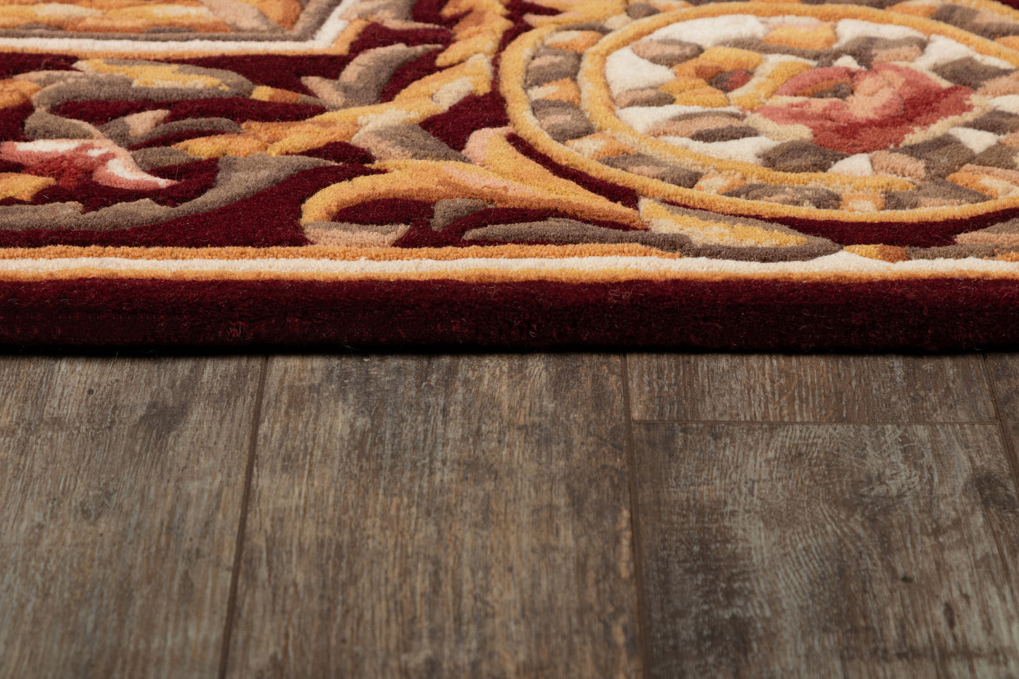 Plush ornate burgundy rug edge with gold, ivory, grey and orange medallion pattern on dark wood flooring.