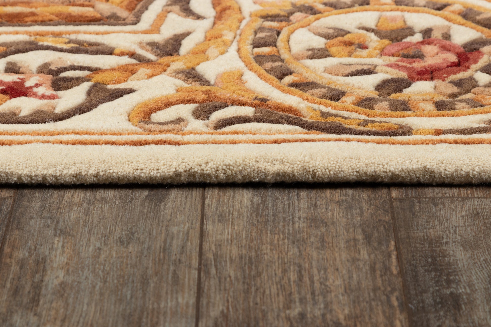 Beige ornate area rug with orange, brown and ivory patterns, plush pile, rolled edge on dark wood floor.