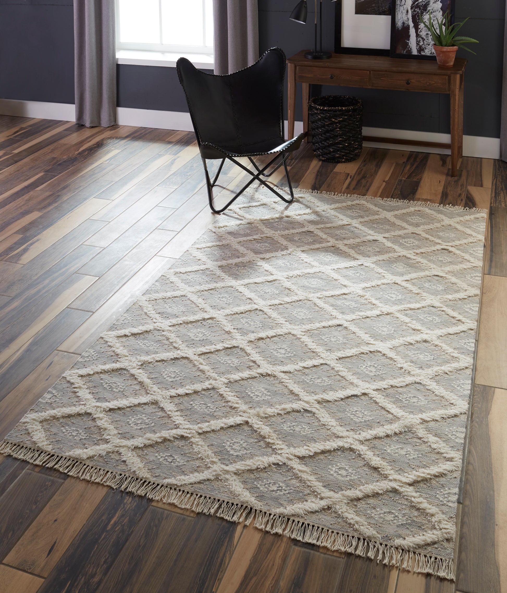 Gray geometric rug with diamond lattice and fringed edges on a dark wood floor, a black butterfly chair and a rustic wood console