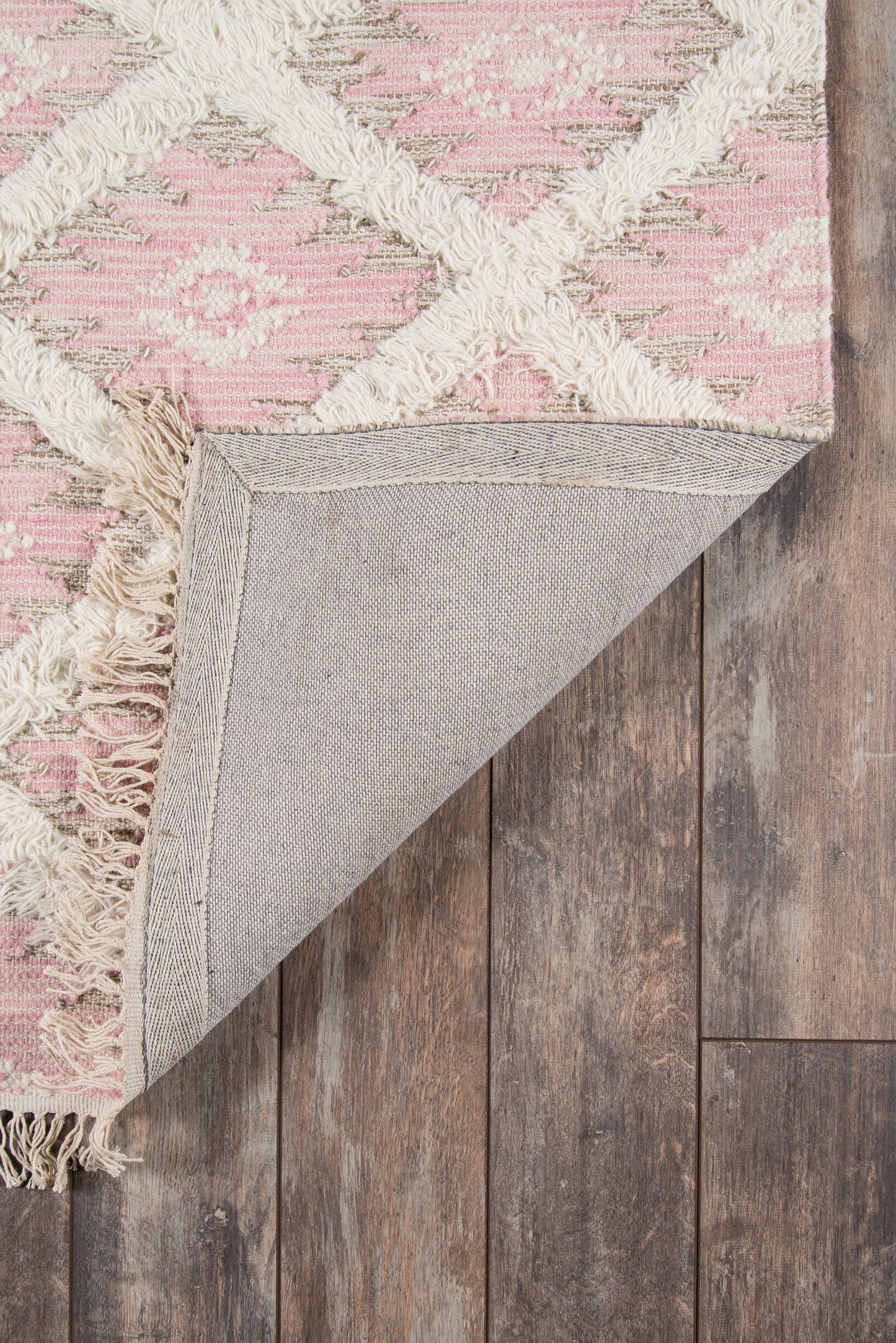 Pink geometric rug with ivory tassels and woven motif; folded corner reveals grey backing on wood floor.