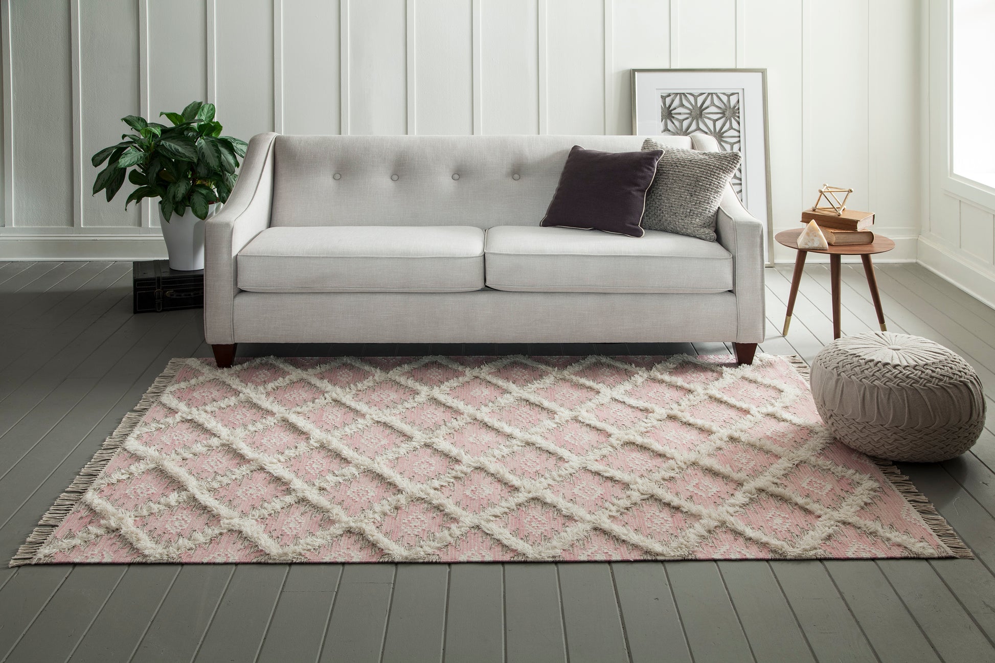 Pink geometric area rug in a bright living room with a light gray tufted sofa, a round knitted pouf, and a plant.