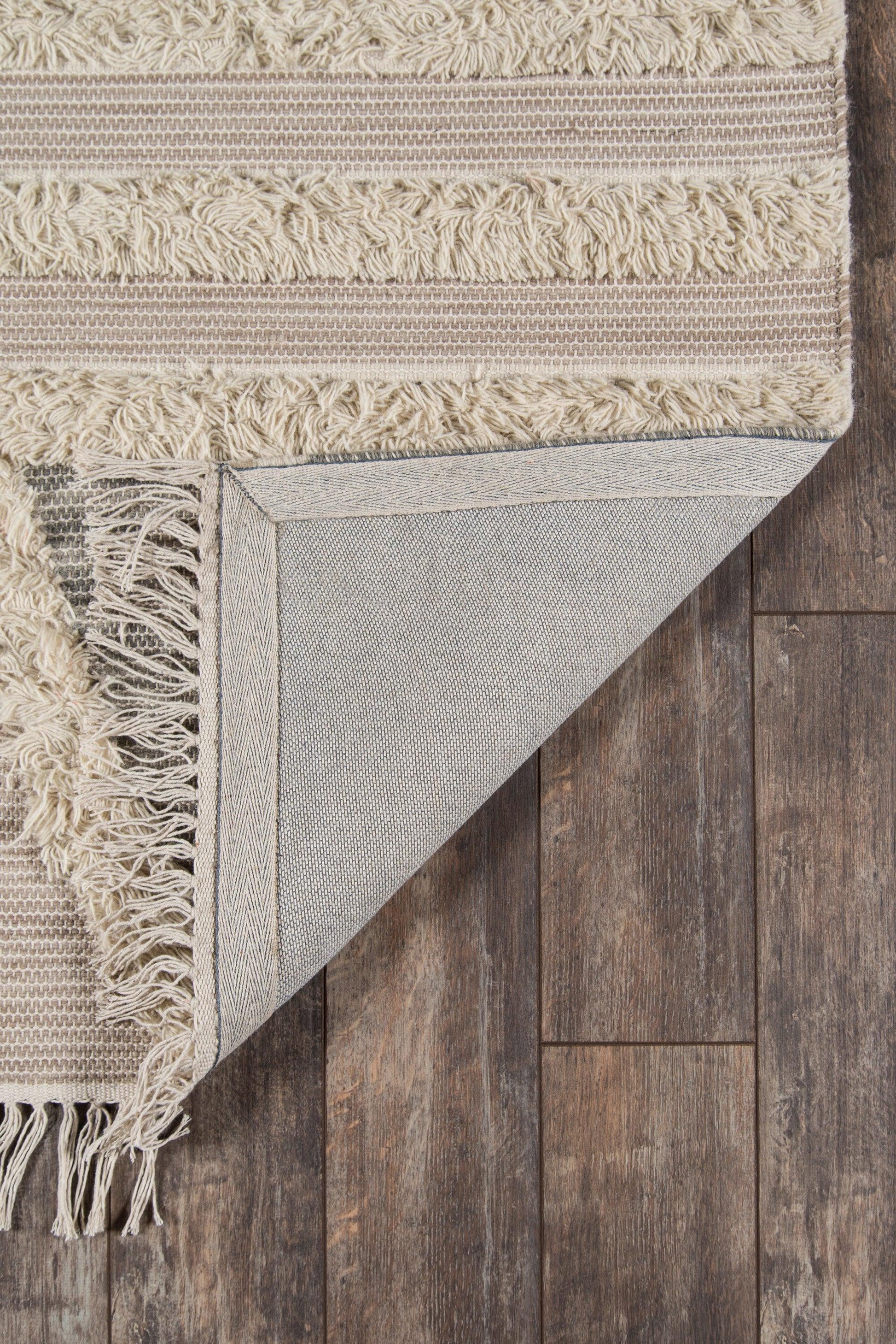 Beige textured rug with striped bands and fringe, folded corner reveals gray backing on dark wood floor.