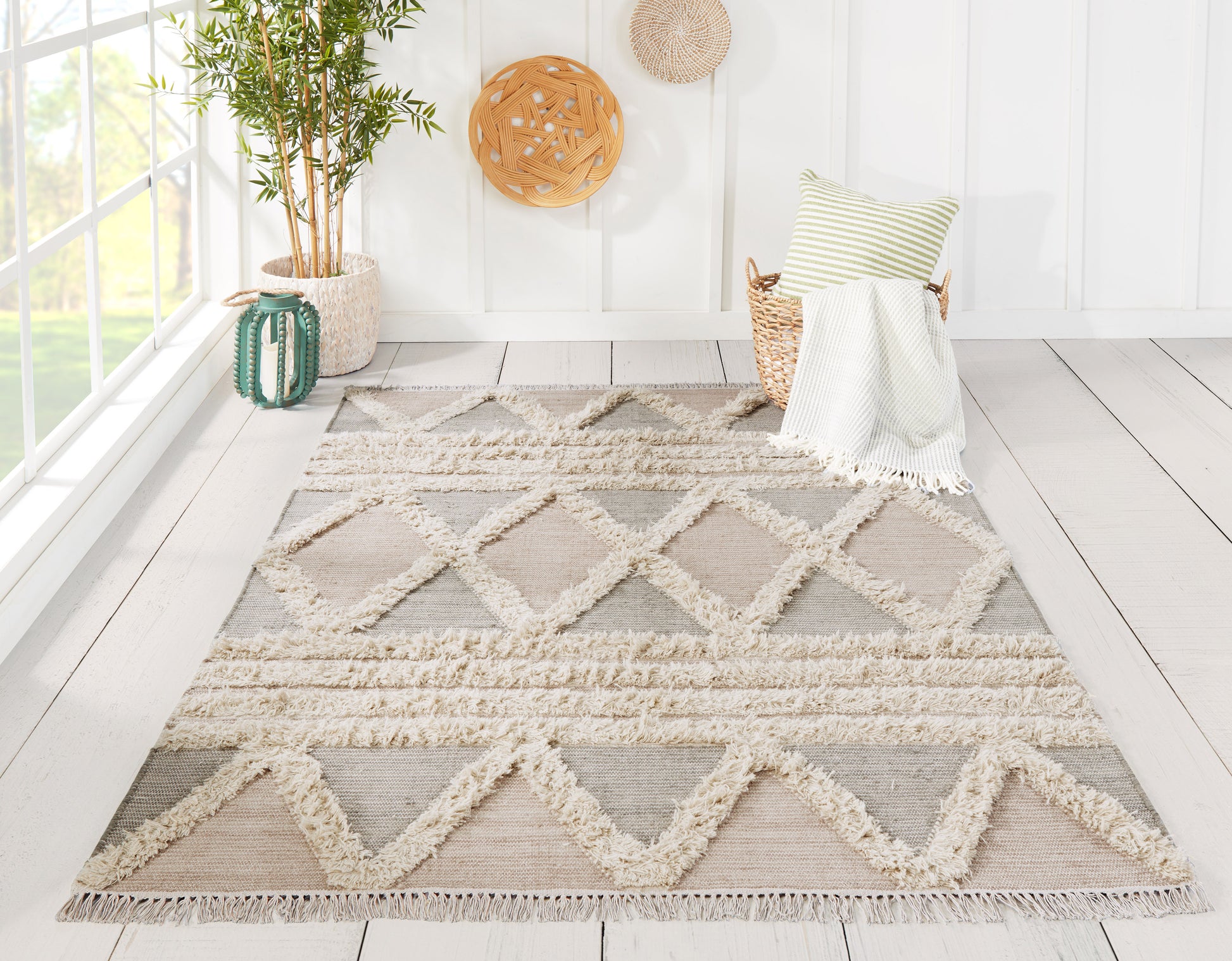 Geometric beige-gray rug with thick cream fringe in a bright sunlit room; wicker chair with striped cushion and cozy throw.