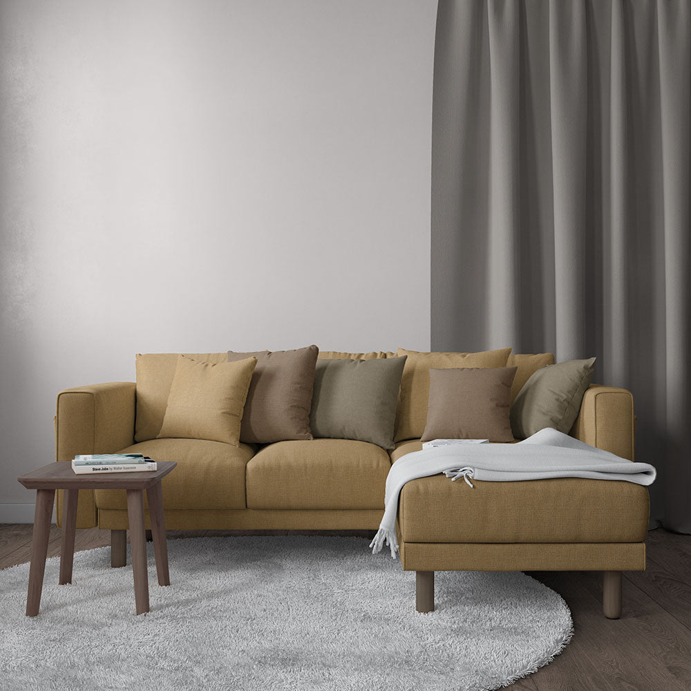 Camel-colored sofa with chaise, assorted beige/olive cushions, light gray throw, wooden side table, gray rug and curtain backdrop.