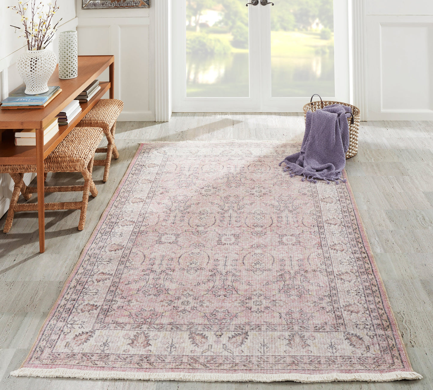 Soft pink oriental area rug with a faded floral border and fringe in a bright living room with wicker stools.