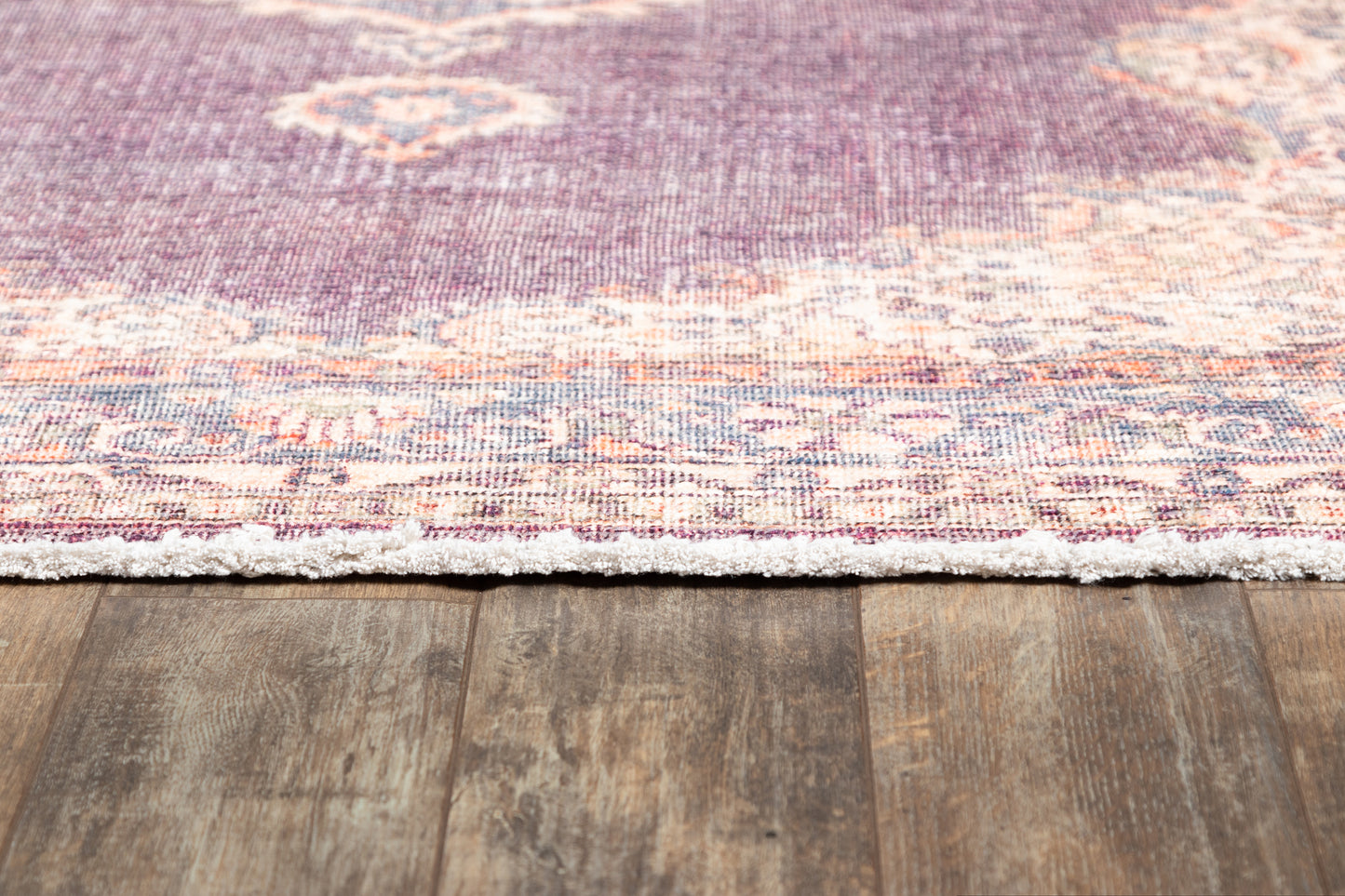 Purple vintage-style rug with faded floral pattern and fringe edge on rustic wood floor.