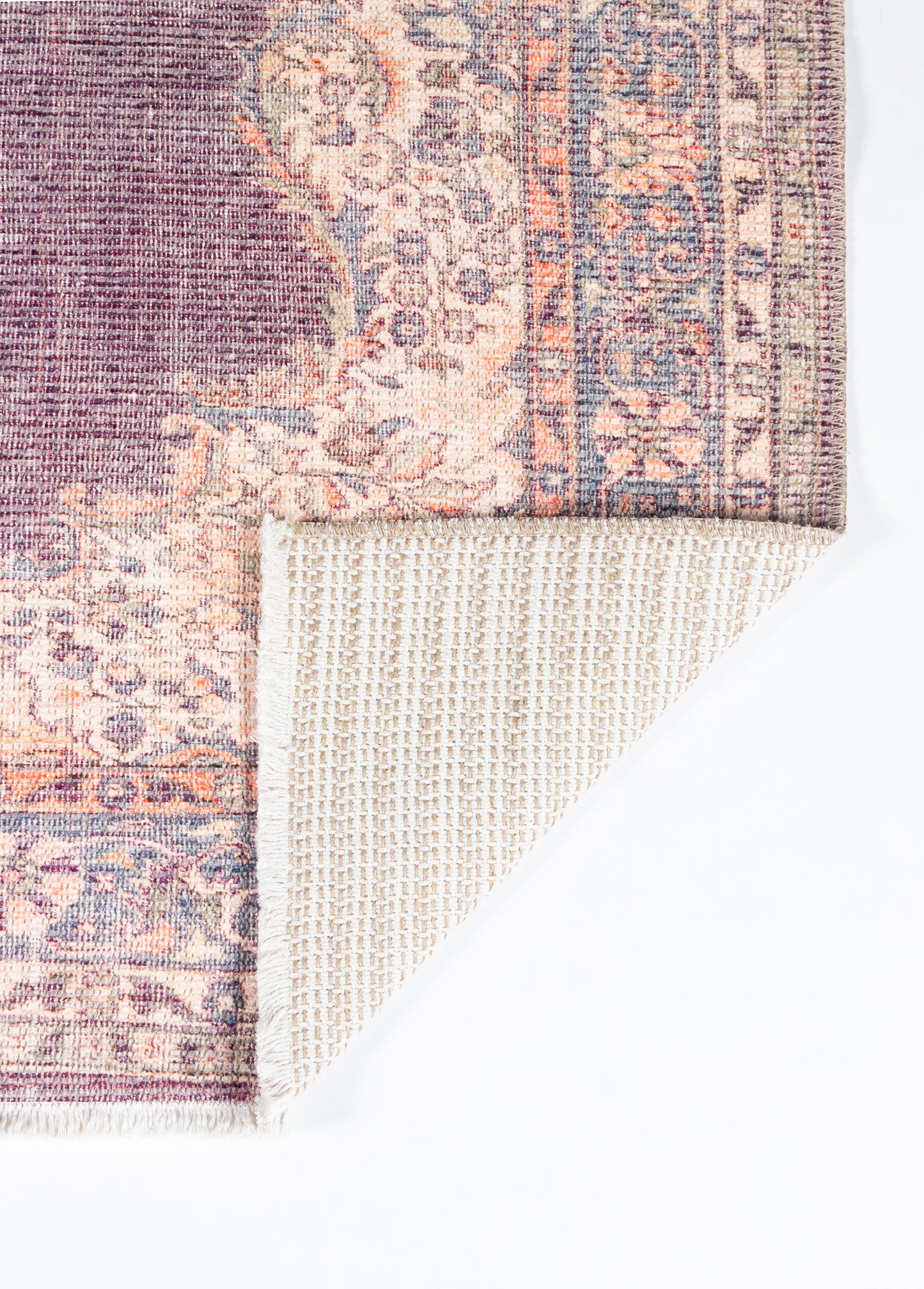 Purple and peach oriental rug with floral border and fringe; folded corner reveals beige grid backing.