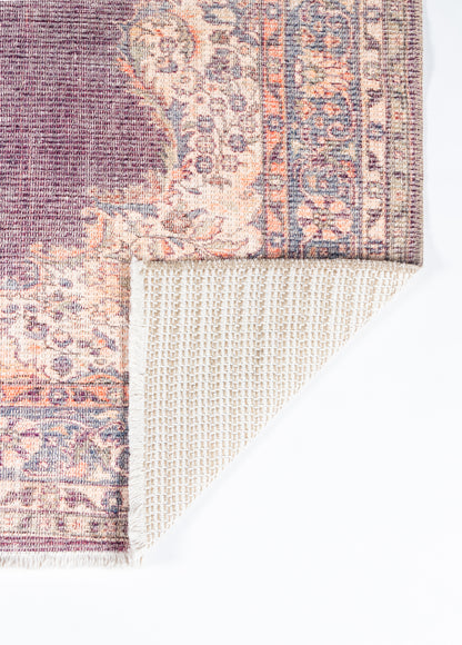 Purple and peach oriental rug with floral border and fringe; folded corner reveals beige grid backing.