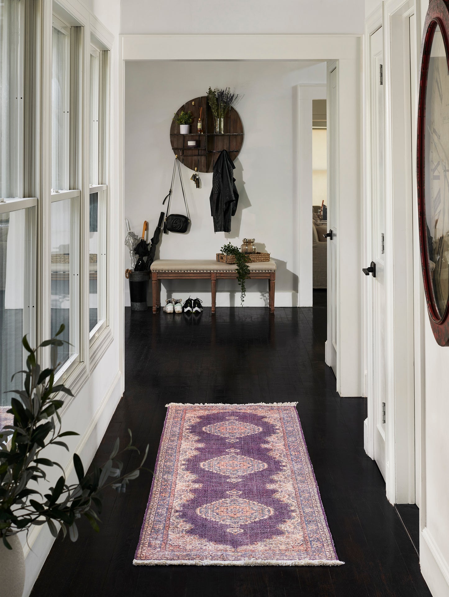 Hallway with purple geometric runner rug, dark wood floor, white walls, bench under circular wooden shelf and coats.