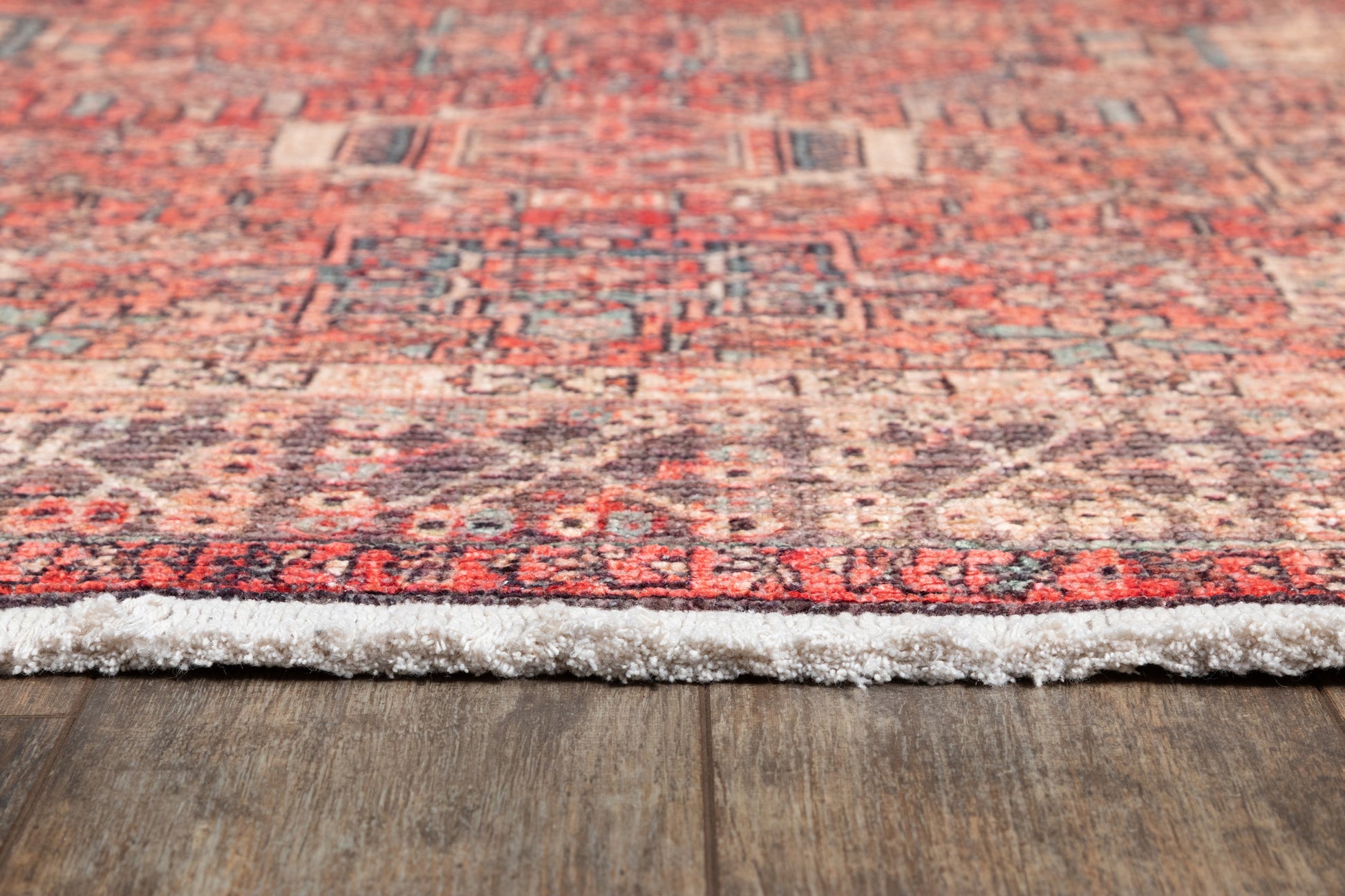 Close-up of a red oriental rug with geometric motifs and white fringe on a wood floor.