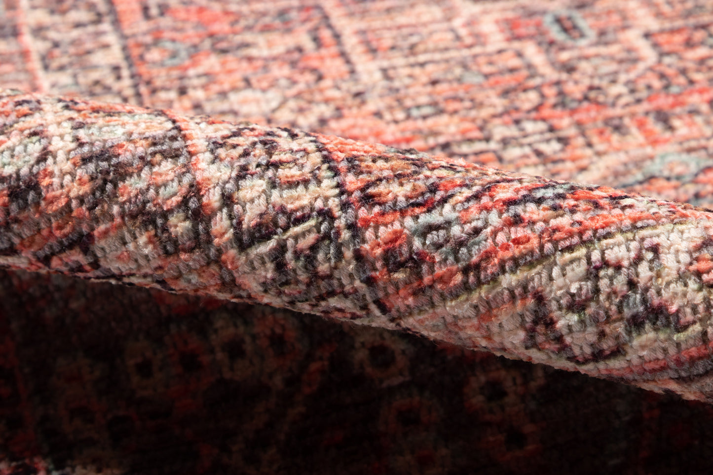 Close-up of a patterned woven rug with red, pink, cream and gray tones; folded corner shows plush pile.