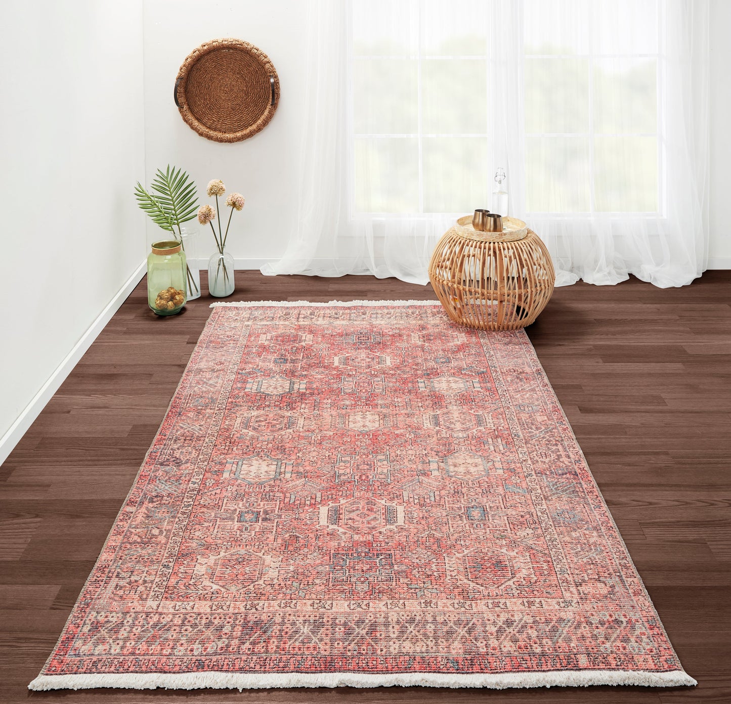 Pink Persian-style rug with geometric medallions on dark wood floor; wicker lantern and glass jars in airy room.