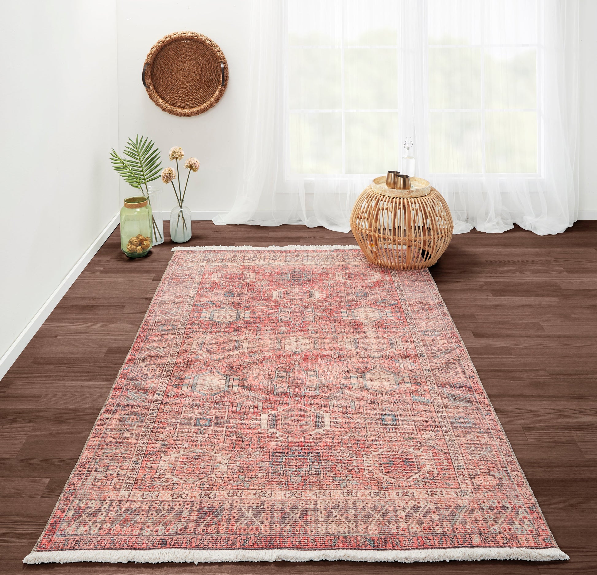 Pink Persian-style rug with geometric medallions on dark wood floor; wicker lantern and glass jars in airy room.