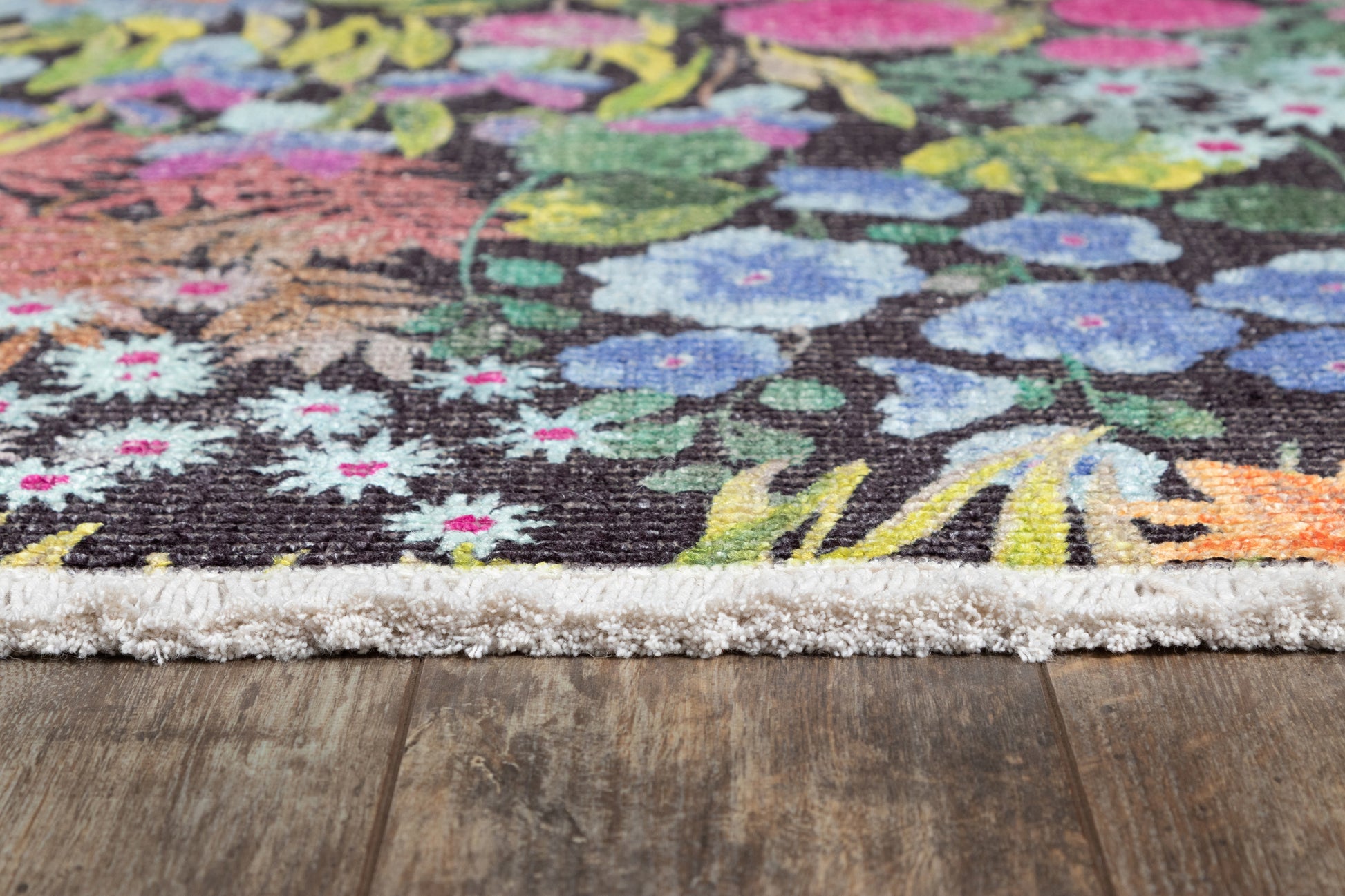 Colorful floral rug edge with white fringe on dark wood floor; woven textile with blue, pink, yellow flowers.