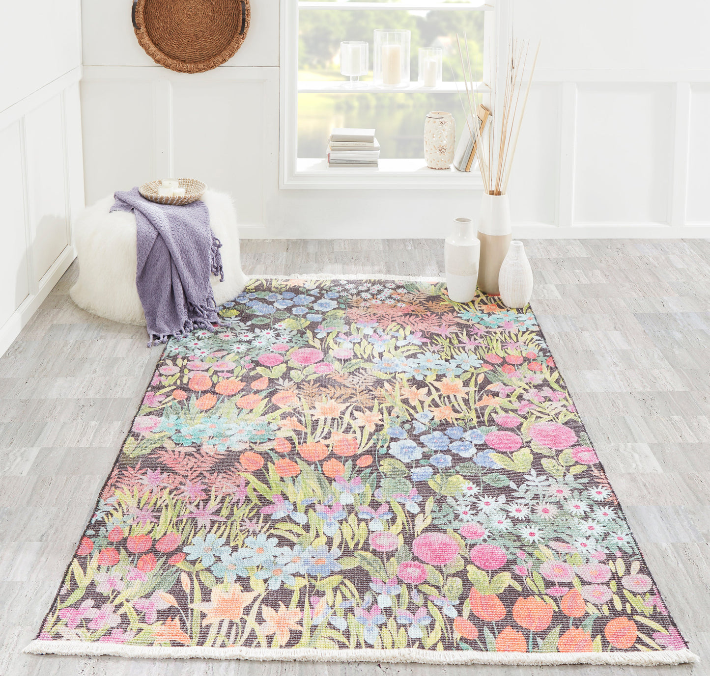 Colorful floral rug on gray wood floor with purple throw on white fluffy pouf, white vases and sunny window.