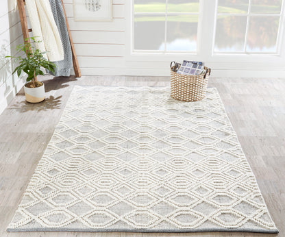 Gray and ivory diamond-patterned woven area rug in a bright, airy room with a potted plant and a woven basket.