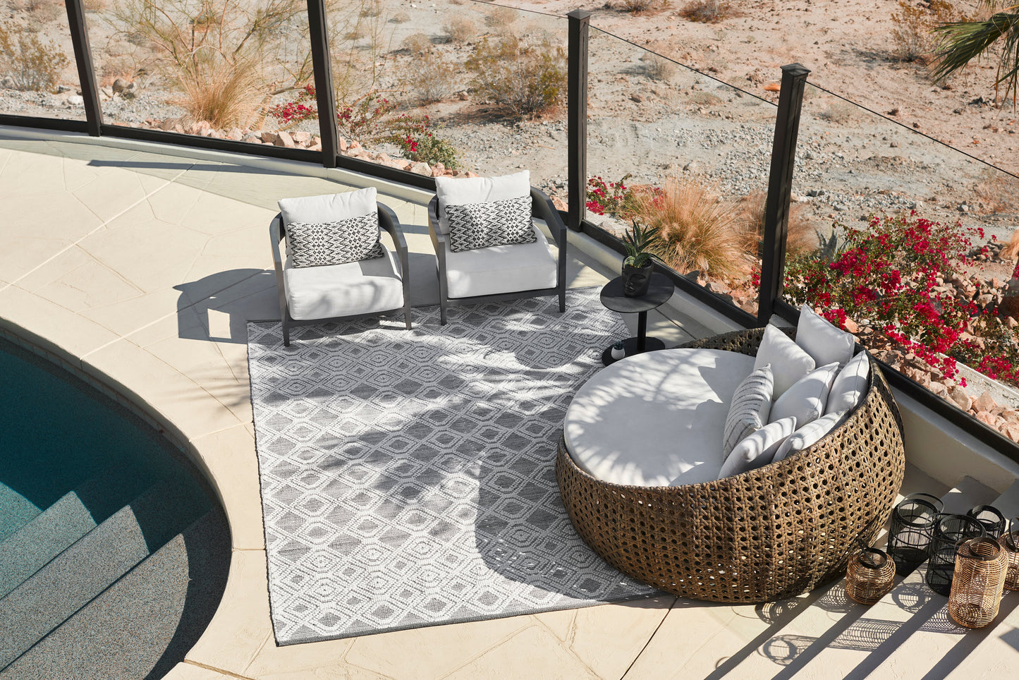 Outdoor lounge set on a gray geometric rug: round wicker daybed with white cushions, two gray-framed chairs, black round side table.