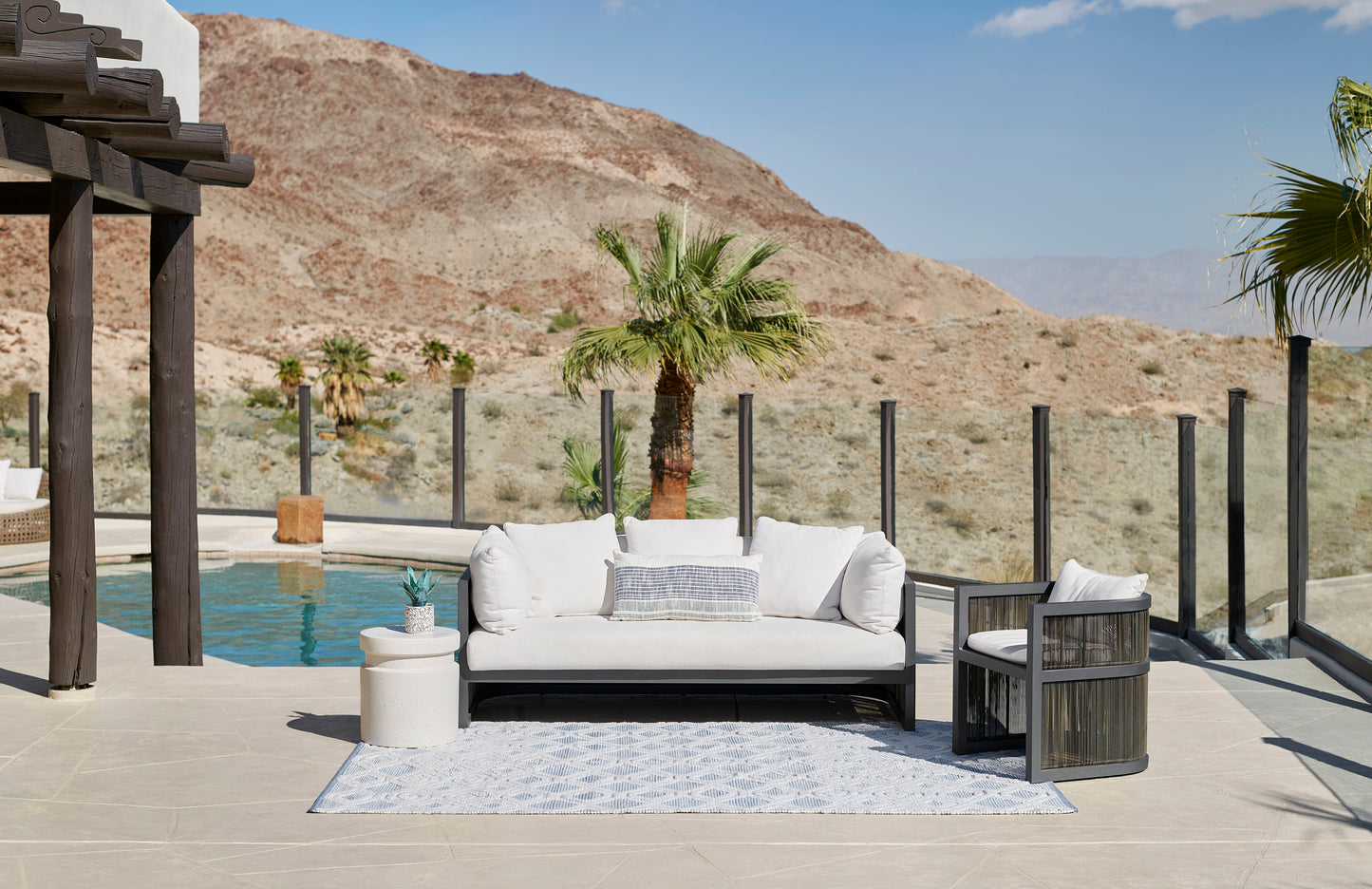 Outdoor lounge set with white cushions on black metal frame, beige-tiled patio, blue patterned rug, pool and desert mountains