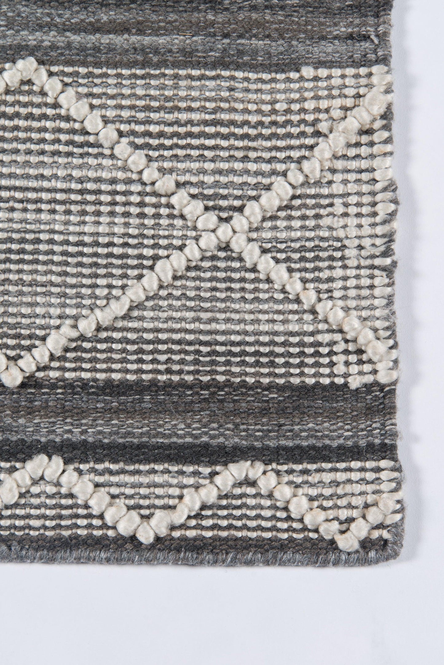 Gray woven rug with cream pom-pom trim creating X-shaped diagonals and zigzag patterns; rustic boho style.