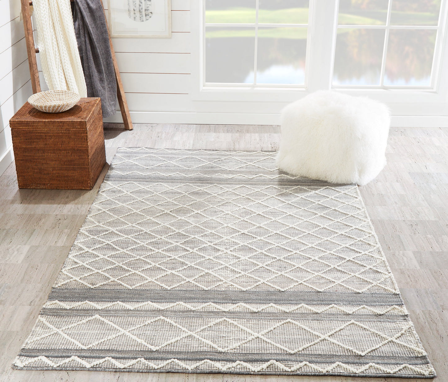 Gray diamond-lattice area rug with textured borders in a bright room, paired with a wicker ottoman and fluffy white pouf.