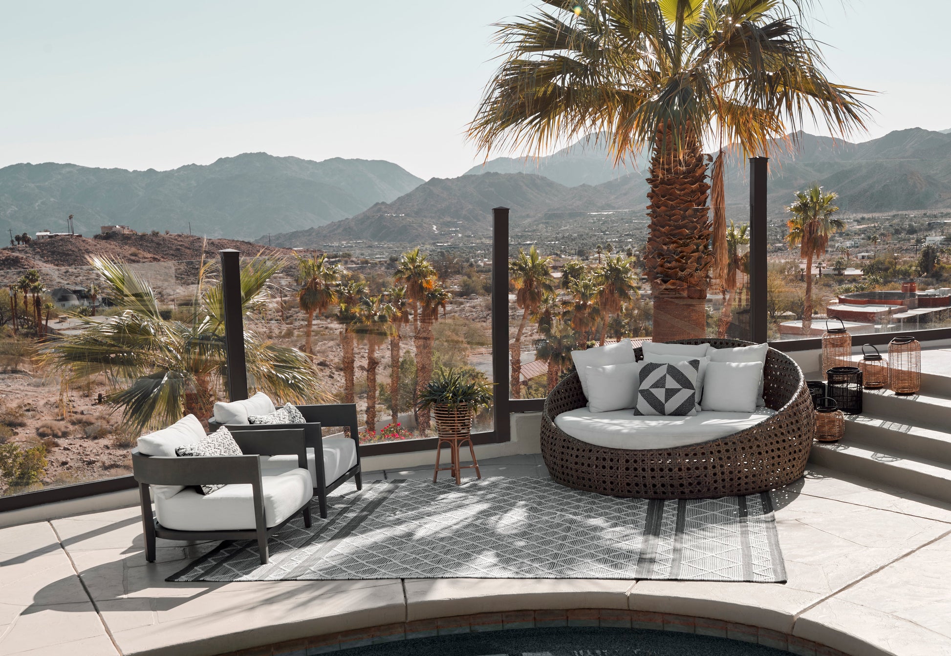 Outdoor patio rug with a geometric pattern beneath round woven rattan seating with white cushions and gray-framed chairs.