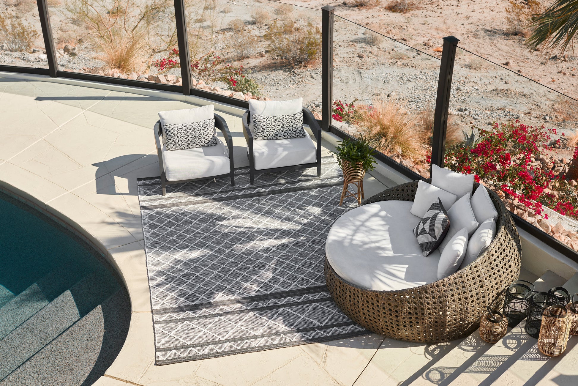 Gray geometric outdoor rug in a seating area with two cushioned armchairs, a round wicker daybed, and poolside glass railing.