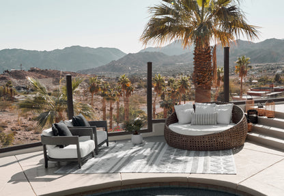 Outdoor striped rug (5' x 8') with a round brown wicker sofa and white cushions, plus two gray armchairs, with desert mountains and palm trees in the background.