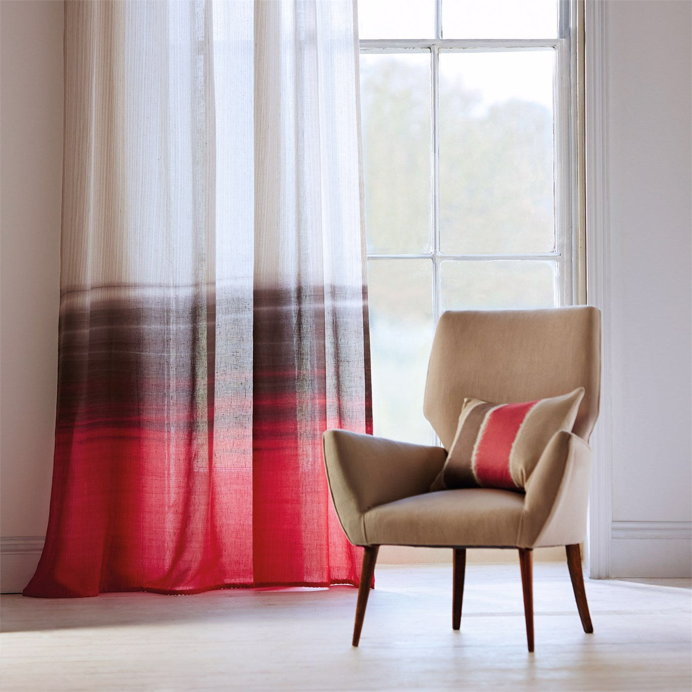 Beige mid-century fabric armchair with wooden legs and a red-striped pillow beside sheer white-to-red gradient curtains.