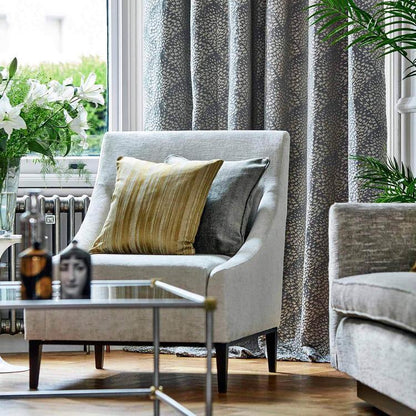 Light gray upholstered armchair with yellow cushion & gray pillow, glass coffee table, metal frame, curtains & plants.