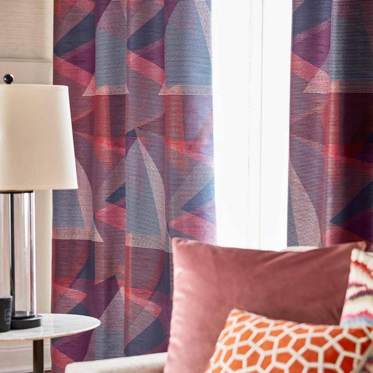 Geometric purple-pink curtains, beige lampshade, round white side table, and pink sofa with an orange geometric cushion.