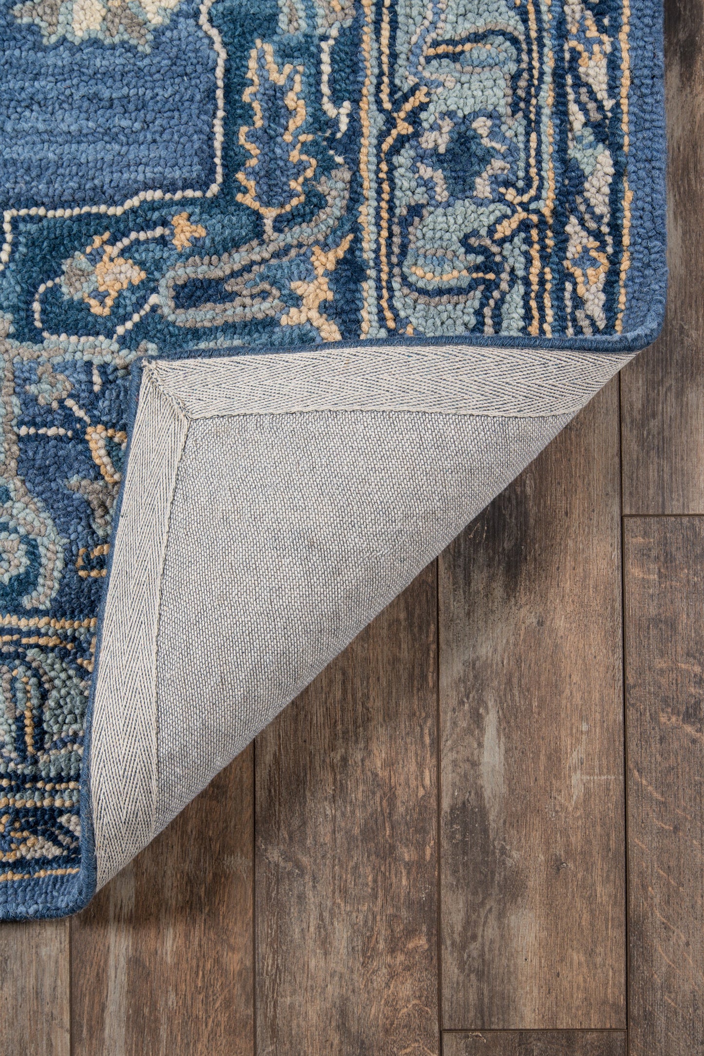 Blue patterned runner rug with ornate gold accents; corner folded to reveal light gray herringbone underside on a wood floor.
