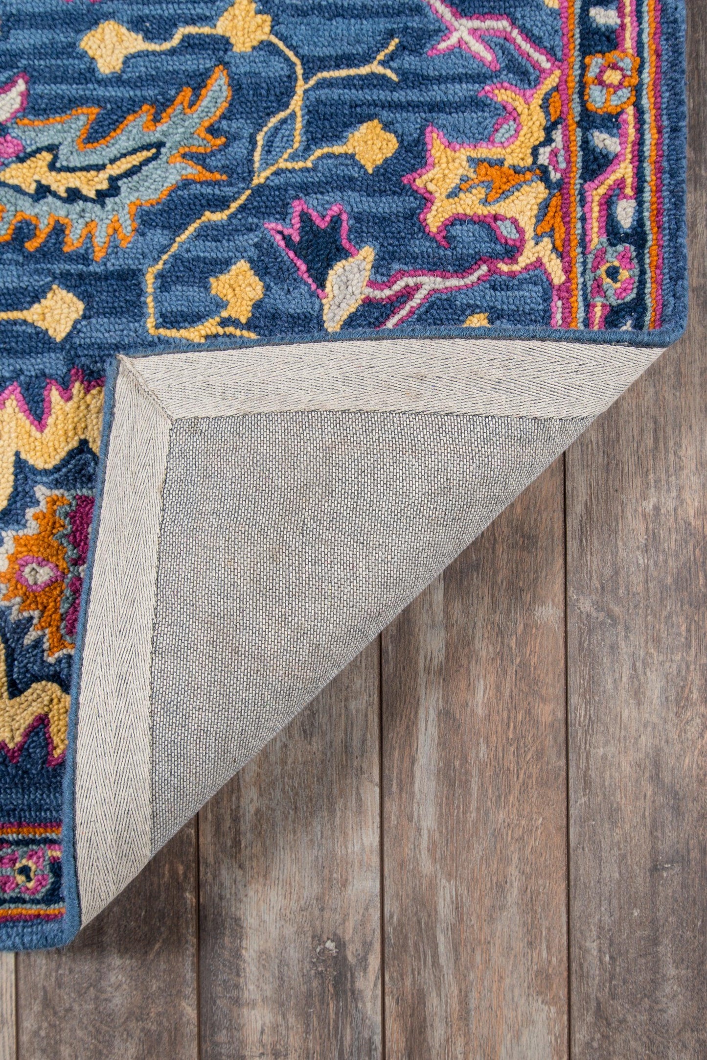 Blue Ibiza rug with pink, orange, yellow motif, thick pile, bordered edge; folded corner shows gray backing on wood floor.