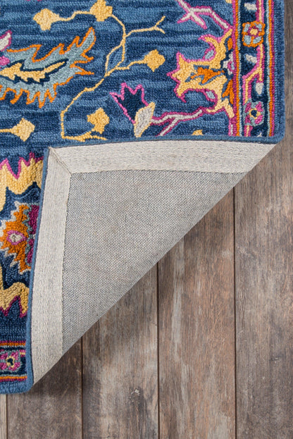 Blue Ibiza rug with pink, orange, yellow motif, thick pile, bordered edge; folded corner shows gray backing on wood floor.