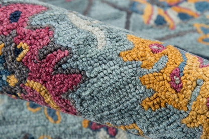 Close-up of a rolled blue-gray rug with pink and mustard floral motifs and teal accents; thick textured pile.