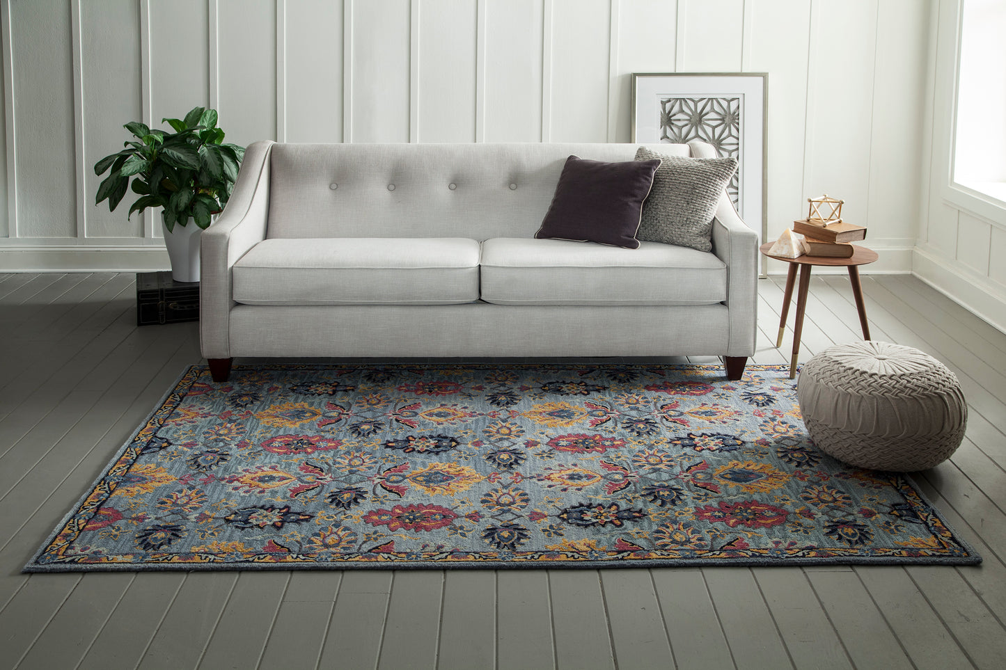 Blue floral area rug with a light gray tufted sofa on it; beige pouf, round wood side table, plant, and framed art.