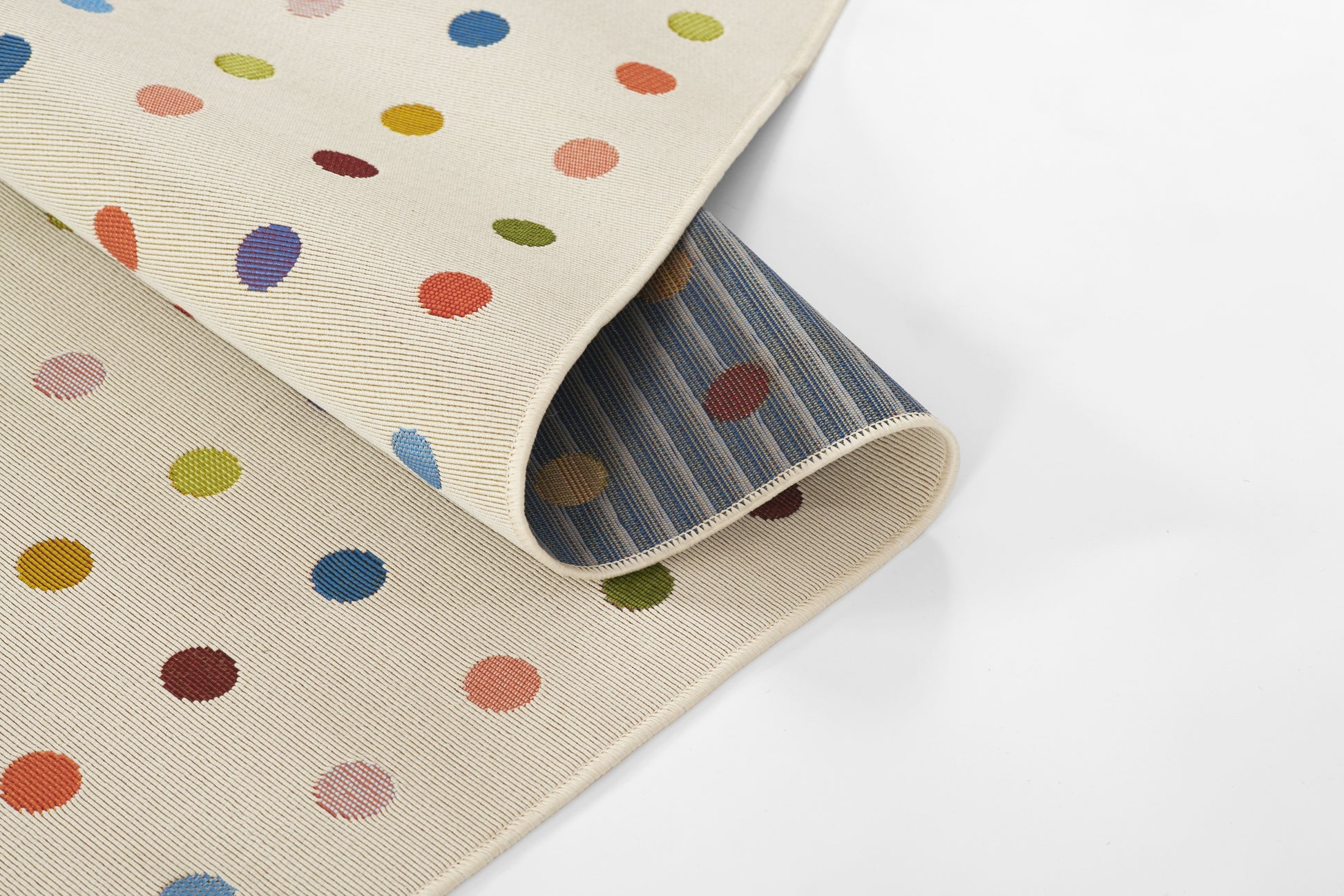 Beige woven rug with multicolor polka dots; rolled to reveal blue striped underside, casual home decor.