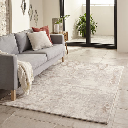 Grey fabric sofa with cream throw & beige cushions on a pale patterned rug; wooden legs, plant & glass balcony door.