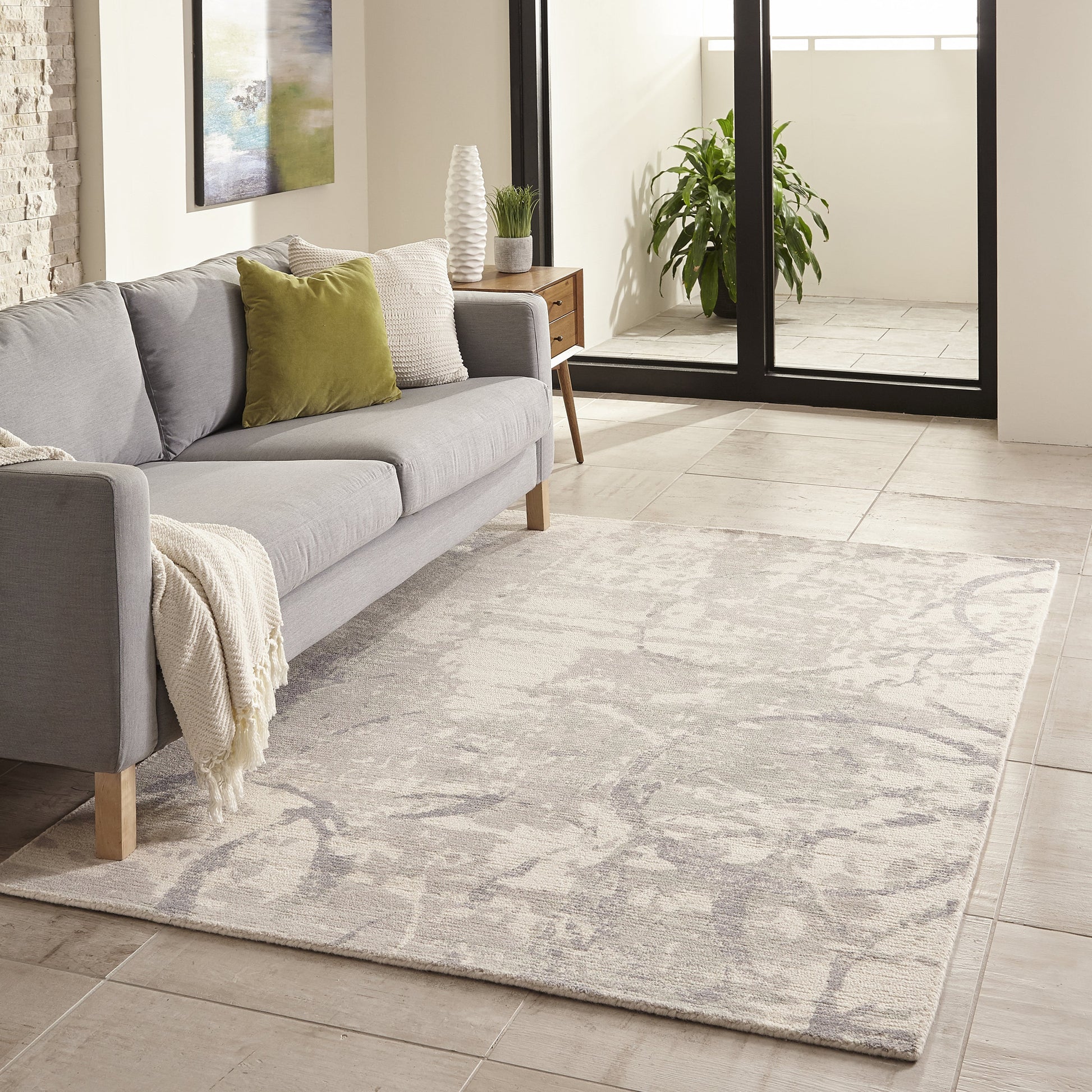 Gray patterned rug in a bright living room with a light gray sofa, knit throw, and green & white pillows near glass doors.