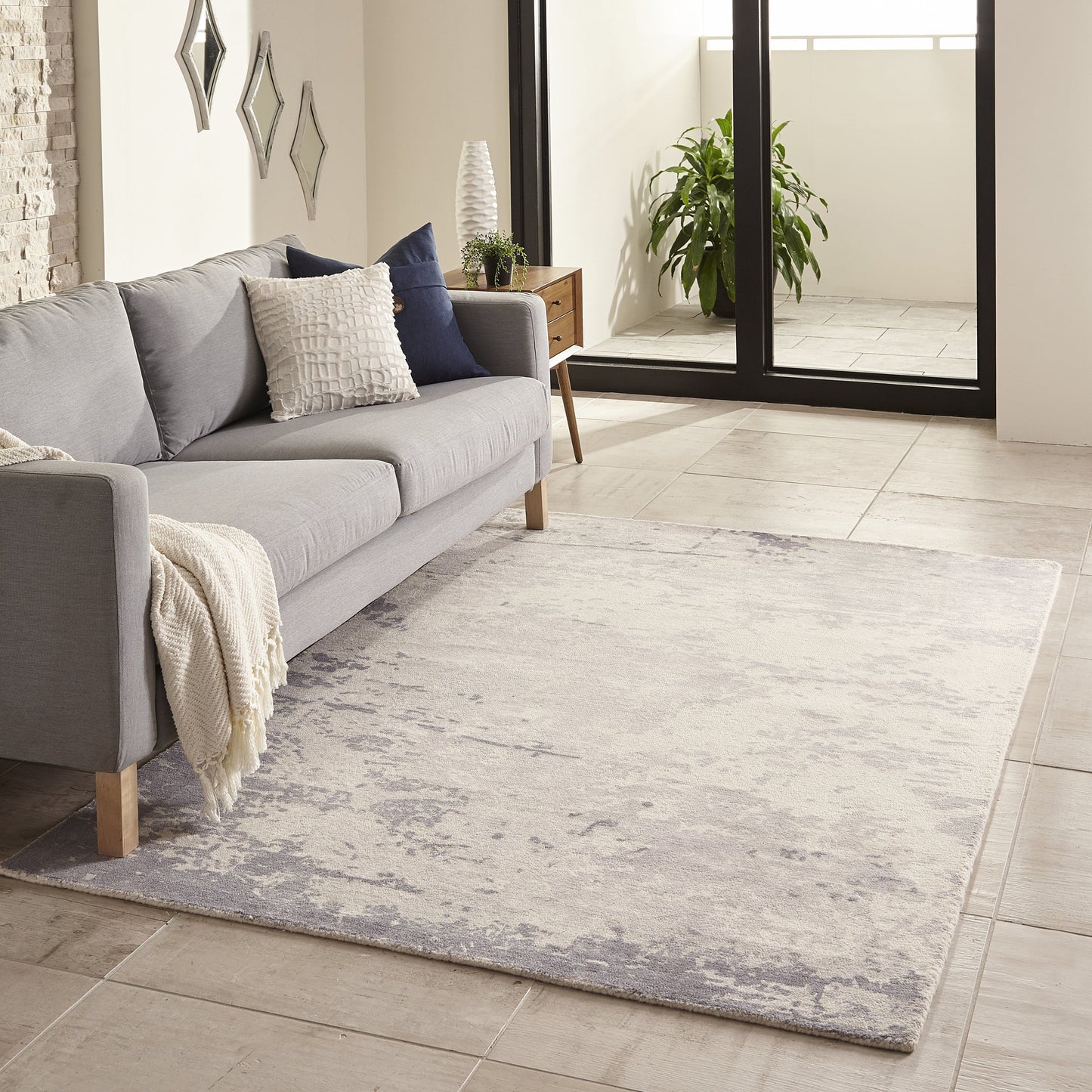 Light gray modern sofa with navy cushions and a cream throw on a pale blue-gray distressed rug in a bright living room.